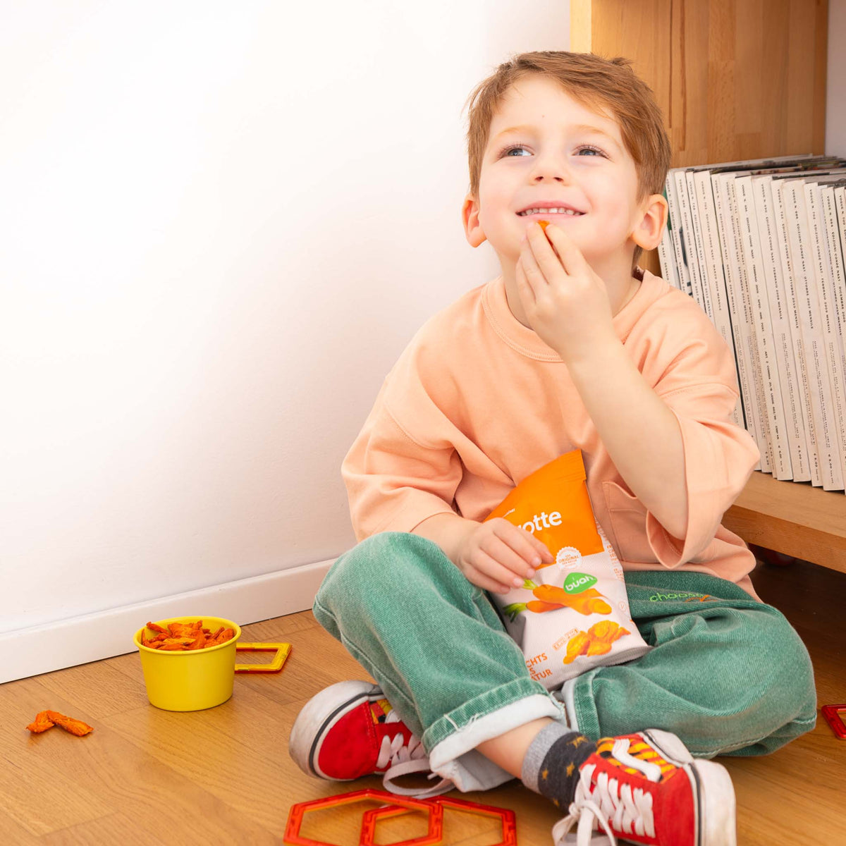 Lachendes Kind sitzt auf dem Boden und isst buah Karottenchips aus der Packung